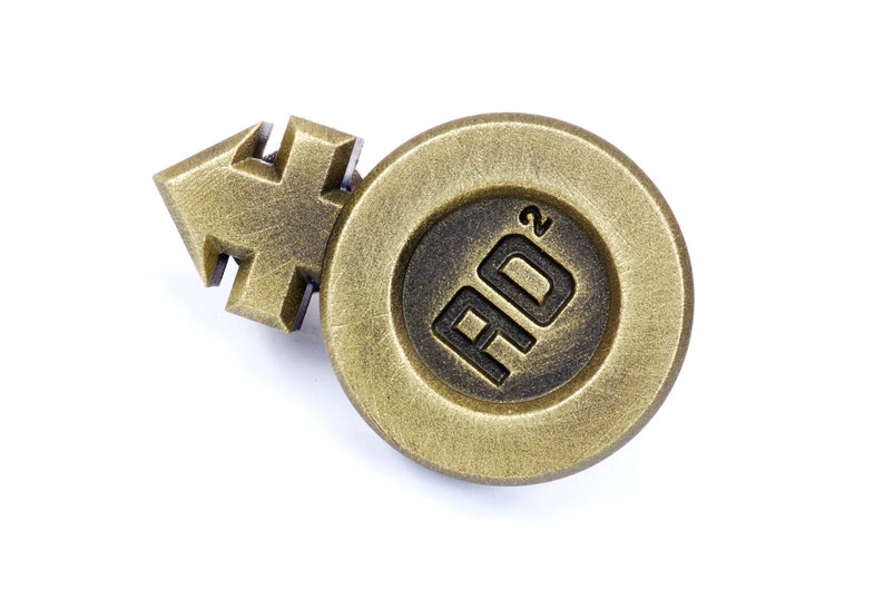 May include: A gold-colored metal pin with a stylized arrow and a circle containing the text "AD&sup2;".