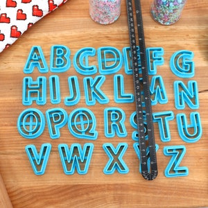 May include: A set of blue plastic alphabet cookie cutters on a wooden surface. The cutters are arranged in alphabetical order.
