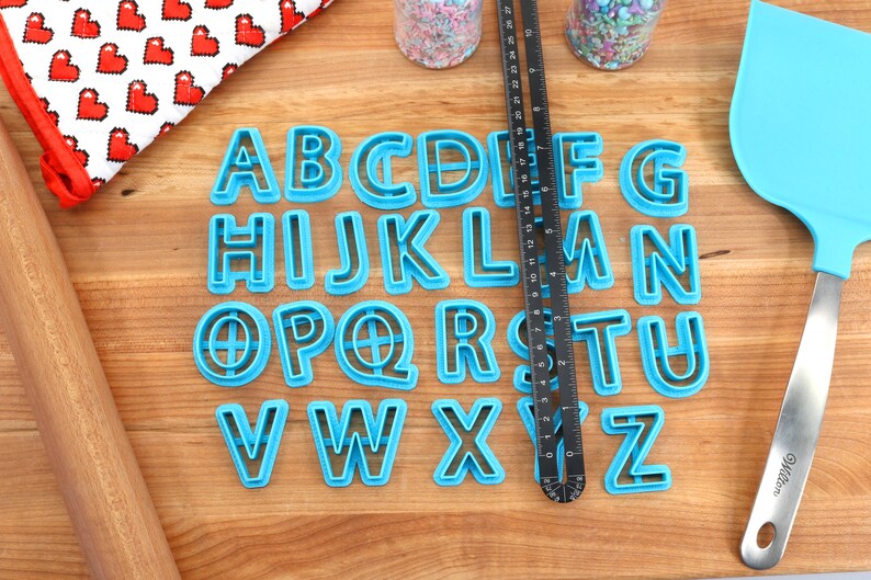 May include: A set of blue plastic alphabet cookie cutters on a wooden surface. The cutters are arranged in alphabetical order.