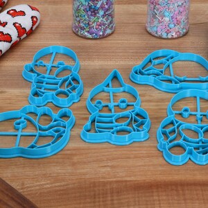 May include: Set of five blue plastic cookie cutters shaped like cartoon characters. The cutters are on a wooden surface.
