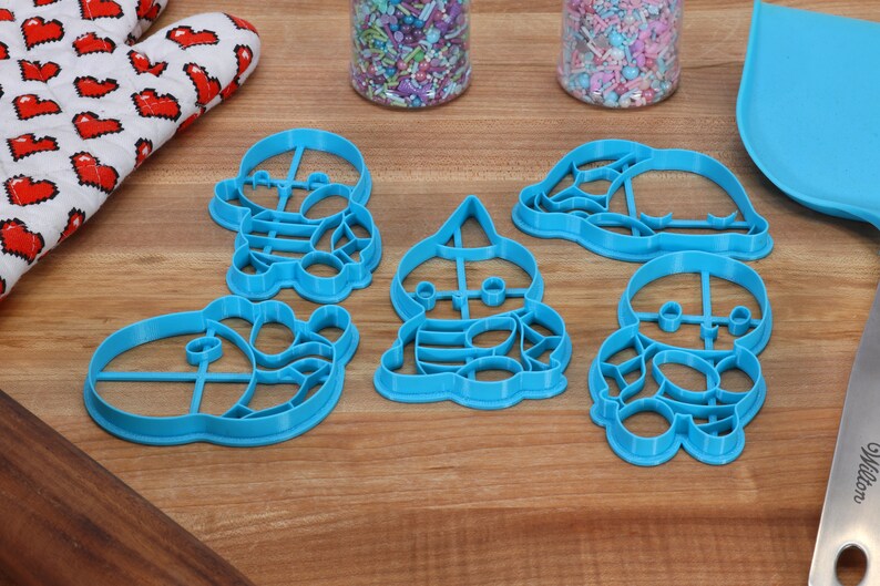 May include: Set of five blue plastic cookie cutters shaped like cartoon characters. The cutters are on a wooden surface.