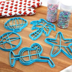 May include: A set of six blue plastic cookie cutters shaped like a seashell, palm tree, beach ball, starfish, cocktail glass, and a slice of citrus. The cutters are on a wooden surface with two jars of colorful sprinkles.