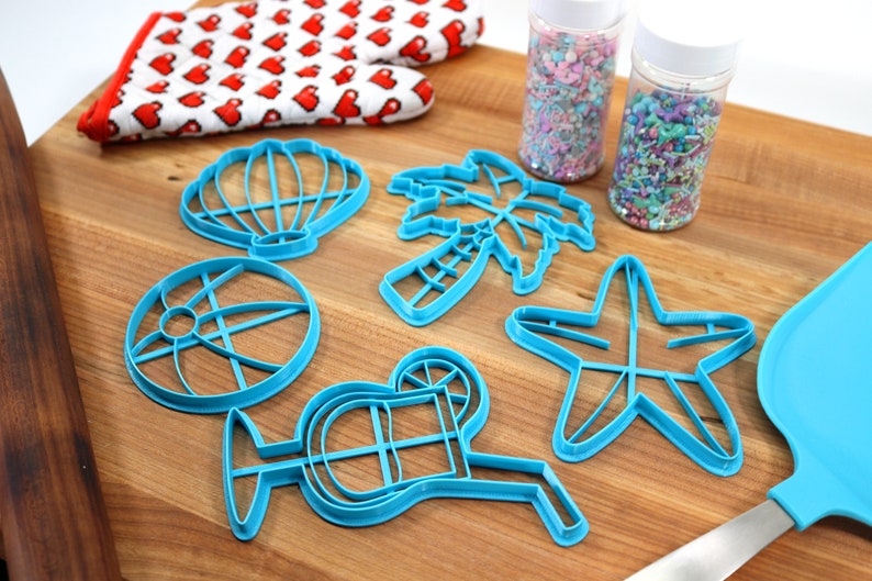 May include: A set of six blue plastic cookie cutters shaped like a seashell, palm tree, beach ball, starfish, cocktail glass, and a slice of citrus. The cutters are on a wooden surface with two jars of colorful sprinkles.