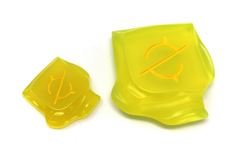 May include: Two translucent yellow resin pieces with a stylized yellow symbol. The larger piece is irregularly shaped, while the smaller one is square. The symbol appears to be a stylized letter or emblem.