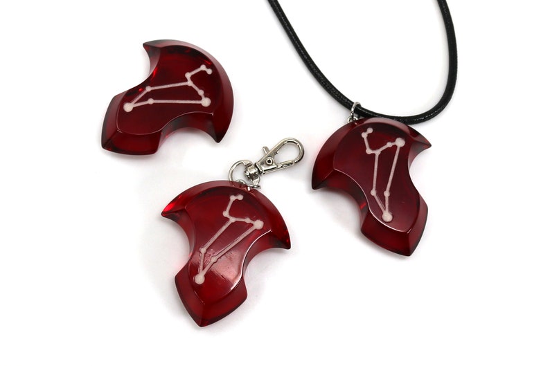 May include: Three red resin pendants with a white constellation design. The pendants are shaped like a crescent moon and have a silver metal ring for hanging. One pendant is on a black cord necklace.