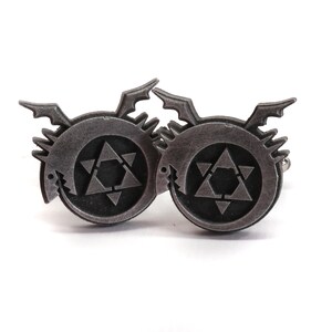 May include: A pair of silver cufflinks featuring the alchemical symbol for transmutation, a circle with a six-pointed star inside, surrounded by a stylized dragon.