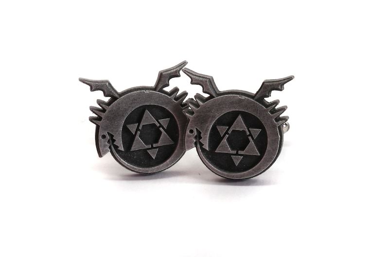 May include: A pair of silver cufflinks featuring the alchemical symbol for transmutation, a circle with a six-pointed star inside, surrounded by a stylized dragon.