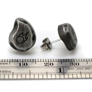 May include: A pair of dark gray, textured stud earrings. Each earring is a rounded, irregular shape with a small, etched design. The earrings are displayed with a metal ruler, showing measurements in inches.