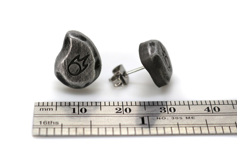 May include: A pair of dark gray, textured stud earrings. Each earring is a rounded, irregular shape with a small, etched design. The earrings are displayed with a metal ruler, showing measurements in inches.