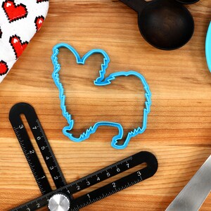 May include: Blue plastic cookie cutter shaped like a Papillon dog on a wooden surface.