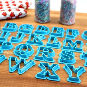 May include: A set of 26 blue plastic alphabet cookie cutters on a wooden surface. The cutters are arranged in alphabetical order.
