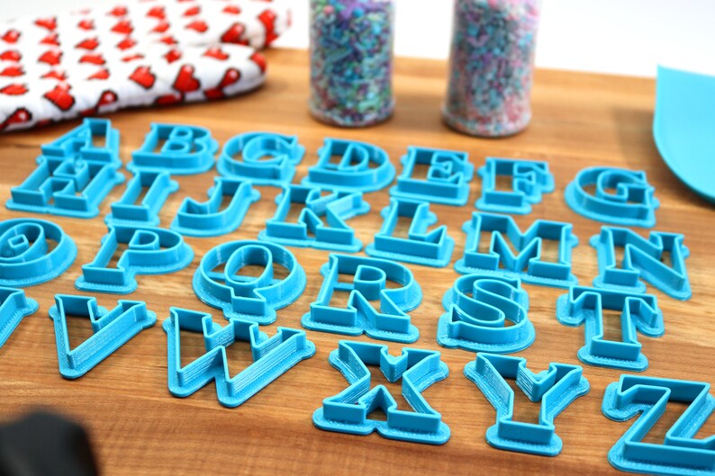 May include: A set of 26 blue plastic alphabet cookie cutters on a wooden surface. The cutters are arranged in alphabetical order.