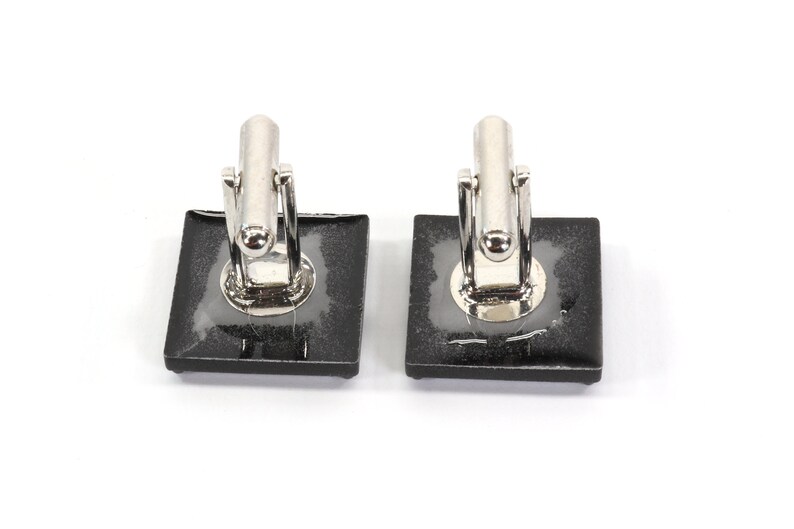 May include: A pair of silver cufflinks with black square-shaped settings. The cufflinks are on a white background.