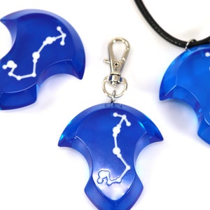 May include: Three blue pendant charms with a white constellation design. The charms are in the shape of a crescent moon with a pointed tip. One charm is on a black cord necklace, one is on a silver chain, and one is loose.