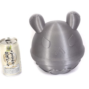 Op de afbeelding: Een grijs 3D-geprint beeldje van een cartoonfiguur met grote oren en een simpel gezicht. Het beeldje staat naast een blikje Japans bier met de tekst "麦芽100%" op het etiket.