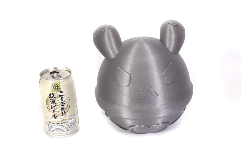 Op de afbeelding: Een grijs 3D-geprint beeldje van een cartoonfiguur met grote oren en een simpel gezicht. Het beeldje staat naast een blikje Japans bier met de tekst "麦芽100%" op het etiket.