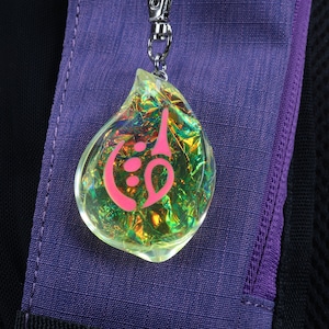 May include: A iridescent yellow teardrop-shaped charm with a pink symbol inside. The charm is attached to a silver chain.