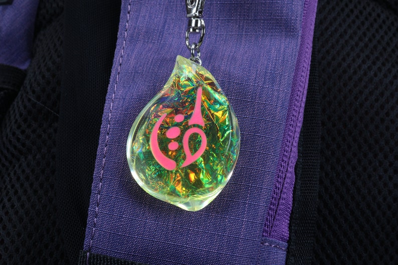 May include: A iridescent yellow teardrop-shaped charm with a pink symbol inside. The charm is attached to a silver chain.