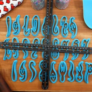 May include: A set of 26 blue plastic cookie cutters shaped like cursive letters of the alphabet. The cutters are arranged on a wooden surface with a ruler and a rolling pin.