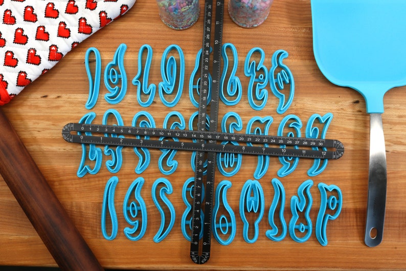 May include: A set of 26 blue plastic cookie cutters shaped like cursive letters of the alphabet. The cutters are arranged on a wooden surface with a ruler and a rolling pin.