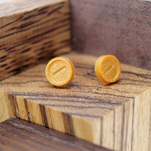 May include: A pair of gold-colored stud earrings with a subtle line design. The earrings are displayed on a wooden surface.