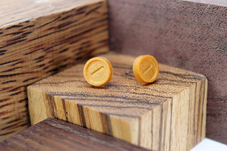 May include: A pair of gold-colored stud earrings with a subtle line design. The earrings are displayed on a wooden surface.