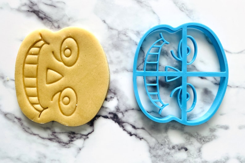 May include: A blue plastic cookie cutter shaped like a skull with a toothy grin and two eye sockets. A plain sugar cookie is shown next to the cutter, which has been cut into the same shape.