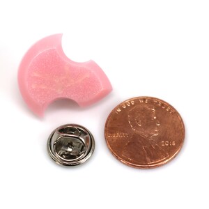 Puede incluir: Un pin de esmalte rosa con forma de media luna con un peque&ntilde;o cierre de pin plateado. El pin es aproximadamente del tama&ntilde;o de una moneda de un centavo de euro.
