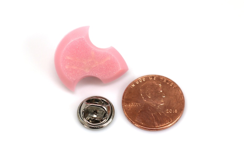 Puede incluir: Un pin de esmalte rosa con forma de media luna con un peque&ntilde;o cierre de pin plateado. El pin es aproximadamente del tama&ntilde;o de una moneda de un centavo de euro.