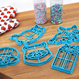 Puede incluir: Cinco cortadores de galletas de pl&aacute;stico azul con forma de mapaches y una TARDIS. Los cortadores est&aacute;n sobre una superficie de madera con dos frascos de chispas en el fondo.