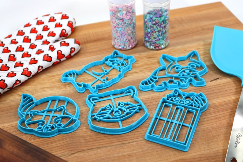 Puede incluir: Cinco cortadores de galletas de pl&aacute;stico azul con forma de mapaches y una TARDIS. Los cortadores est&aacute;n sobre una superficie de madera con dos frascos de chispas en el fondo.