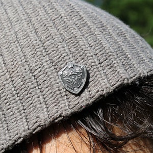 May include: A grey knitted beanie with a silver shield pin featuring a winged design.