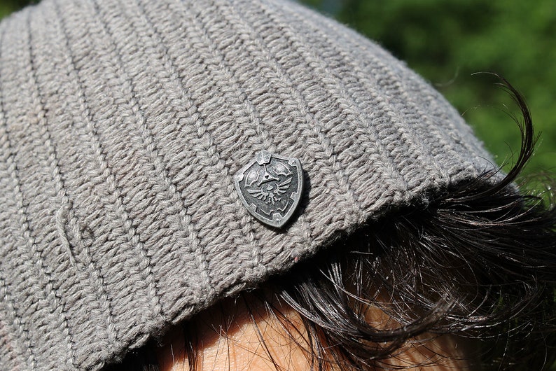 May include: A grey knitted beanie with a silver shield pin featuring a winged design.