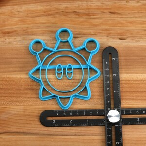 May include: A blue plastic cookie cutter shaped like a cartoon sun with two eyes and a mouth. The cutter is on a wooden surface with a ruler next to it.