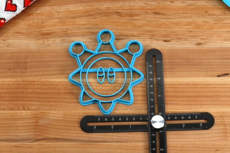 May include: A blue plastic cookie cutter shaped like a cartoon sun with two eyes and a mouth. The cutter is on a wooden surface with a ruler next to it.