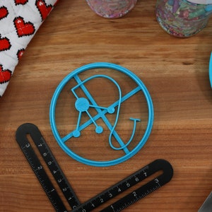 May include: A blue plastic cookie cutter shaped like a stylized letter "A" with a circle in the center and a curved line extending from the top. The cookie cutter is on a wooden surface.