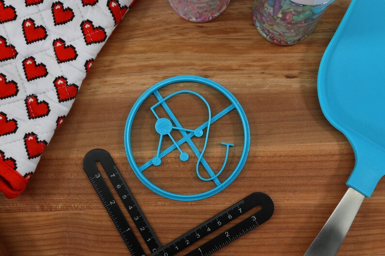 May include: A blue plastic cookie cutter shaped like a stylized letter "A" with a circle in the center and a curved line extending from the top. The cookie cutter is on a wooden surface.