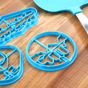 May include: Three blue plastic cookie cutters on a wooden surface. The cutters are shaped like an alien head, a sign that says "Welcome to Area 52", and a UFO.