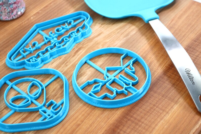 May include: Three blue plastic cookie cutters on a wooden surface. The cutters are shaped like an alien head, a sign that says "Welcome to Area 52", and a UFO.