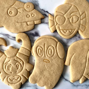 K&ouml;nnte beinhalten: F&uuml;nf Zuckerkekse, die mit verschiedenen Comicfiguren verziert sind. Die Kekse sind hellbraun und haben wei&szlig;e Zuckergussdetails. Zu den Figuren geh&ouml;ren ein Geist, ein Monster mit H&ouml;rnern, eine Kreatur mit einem gro&szlig;en Auge und zwei weitere Figuren mit gro&szlig;en Augen.