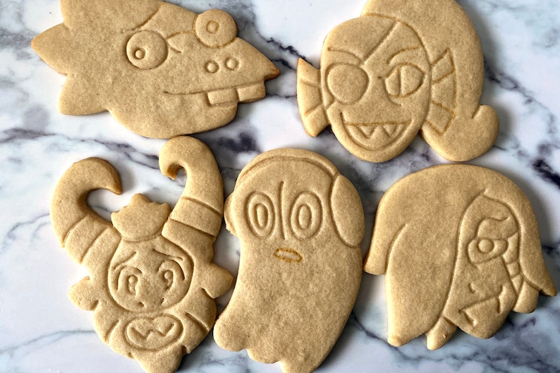 K&ouml;nnte beinhalten: F&uuml;nf Zuckerkekse, die mit verschiedenen Comicfiguren verziert sind. Die Kekse sind hellbraun und haben wei&szlig;e Zuckergussdetails. Zu den Figuren geh&ouml;ren ein Geist, ein Monster mit H&ouml;rnern, eine Kreatur mit einem gro&szlig;en Auge und zwei weitere Figuren mit gro&szlig;en Augen.