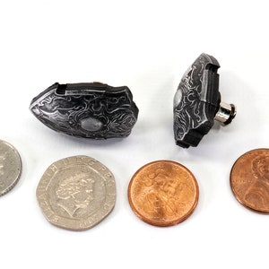 May include: Two small, silver-coloured, ornate, shield-shaped cufflinks. The cufflinks are lying on a white surface next to four coins.