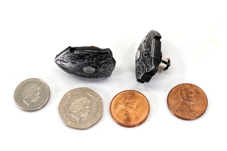 May include: Two small, silver-coloured, ornate, shield-shaped cufflinks. The cufflinks are lying on a white surface next to four coins.