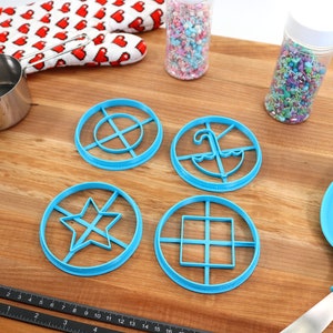 May include: A collection of baking tools and supplies on a wooden surface. Includes blue cookie cutters in various shapes, a rolling pin, measuring cup, oven mitt with heart pattern, sprinkles, and a blue spatula.
