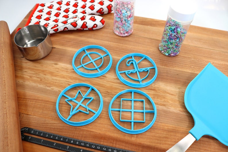 May include: A collection of baking tools and supplies on a wooden surface. Includes blue cookie cutters in various shapes, a rolling pin, measuring cup, oven mitt with heart pattern, sprinkles, and a blue spatula.