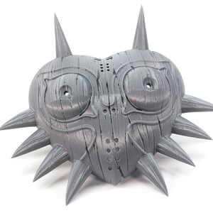 May include: A gray 3D printed mask shaped like a heart with spikes around the edges. The mask has two large eye holes and a smaller hole in the center.