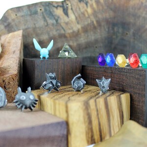 May include: A collection of silver-colored metal rings featuring various designs from the Legend of Zelda video game series. The rings are displayed on small wooden blocks. The designs include a Triforce, a Master Sword, a Hylian Shield, a Deku Scrub, and a Ganondorf.