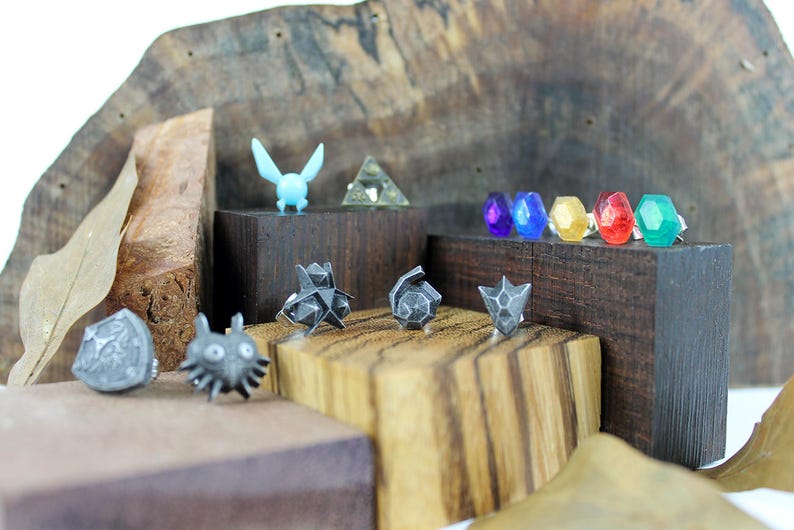 May include: A collection of silver-colored metal rings featuring various designs from the Legend of Zelda video game series. The rings are displayed on small wooden blocks. The designs include a Triforce, a Master Sword, a Hylian Shield, a Deku Scrub, and a Ganondorf.