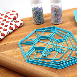 May include: A blue plastic snowflake cookie cutter on a wooden surface with a rolling pin, oven mitts, and two jars of sprinkles.