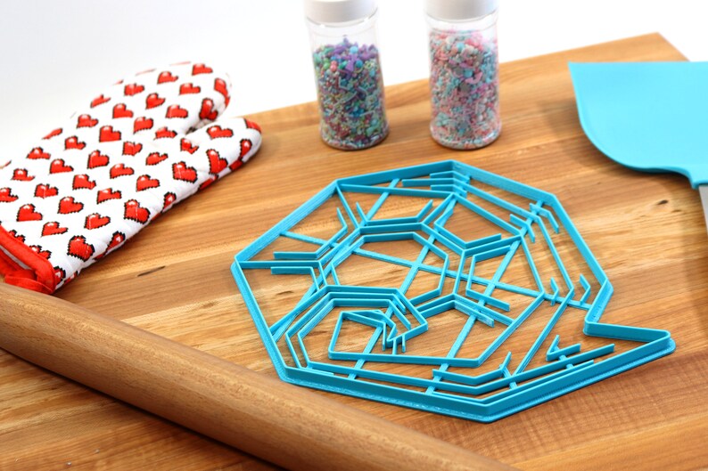May include: A blue plastic snowflake cookie cutter on a wooden surface with a rolling pin, oven mitts, and two jars of sprinkles.